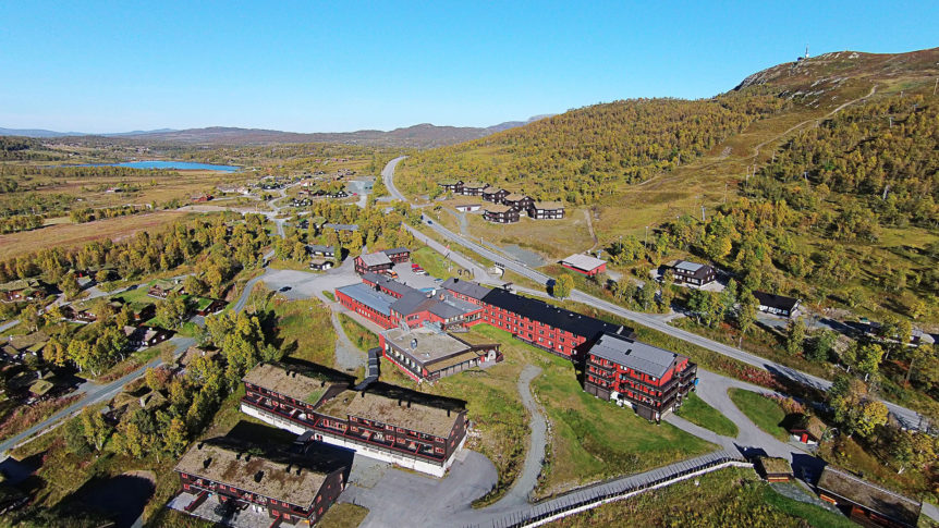 Flyfoto av Røros landskap med røde bygninger og en innsjø i bakgrunnen. Høstfarger dekker området.