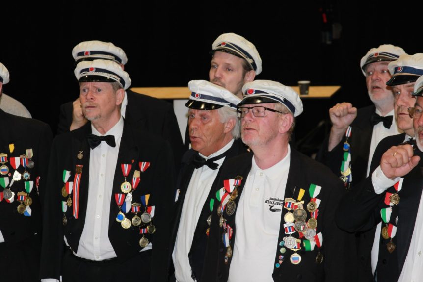 Eldre menn i uniform med medaljer, deltar i en syngende gruppe, bærende tradisjonelle hatter og slips.