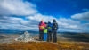 Familie på fjelltur omfavner hverandre med panoramautsikt over landskapet under en blå himmel.