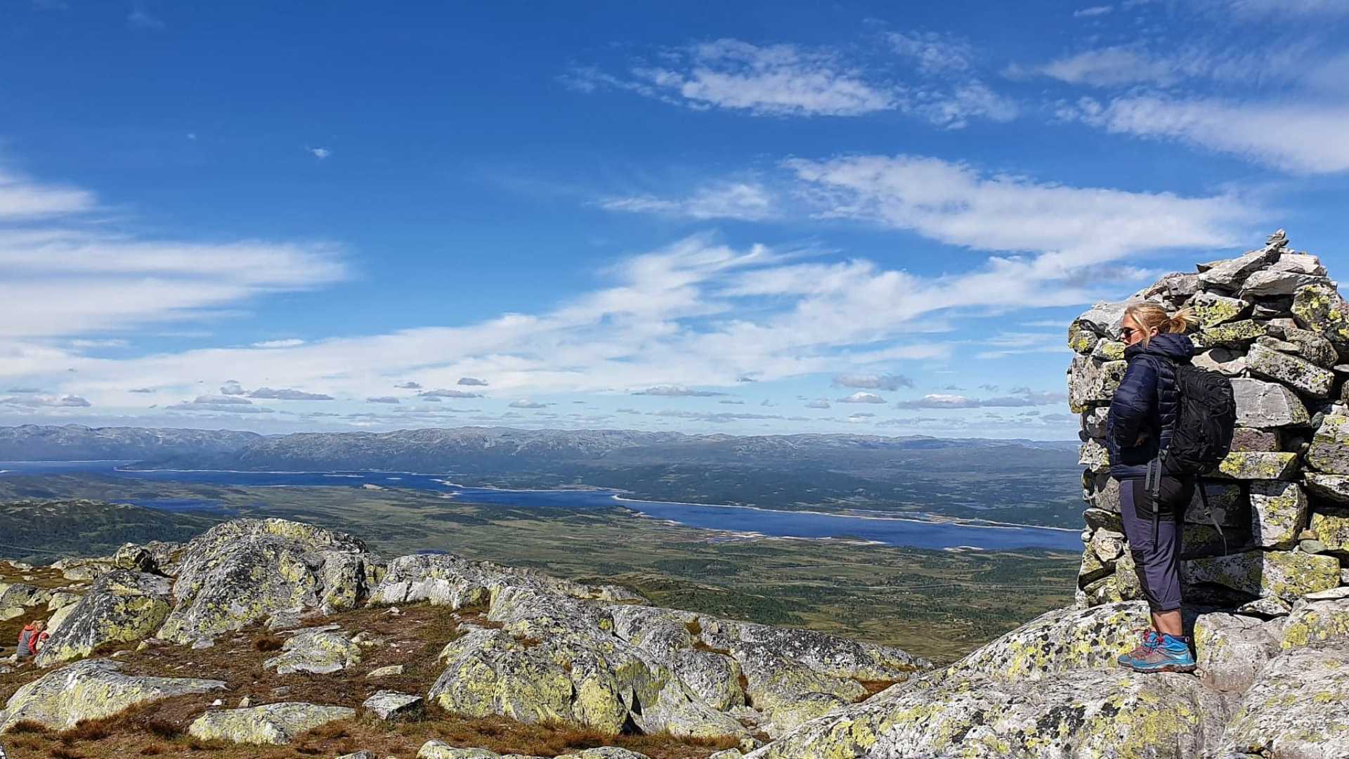 Kvinne på fjelltur i fjellandskap, steinmur ved siden av henne, blå himmel og vidstrakt utsikt over dalen.