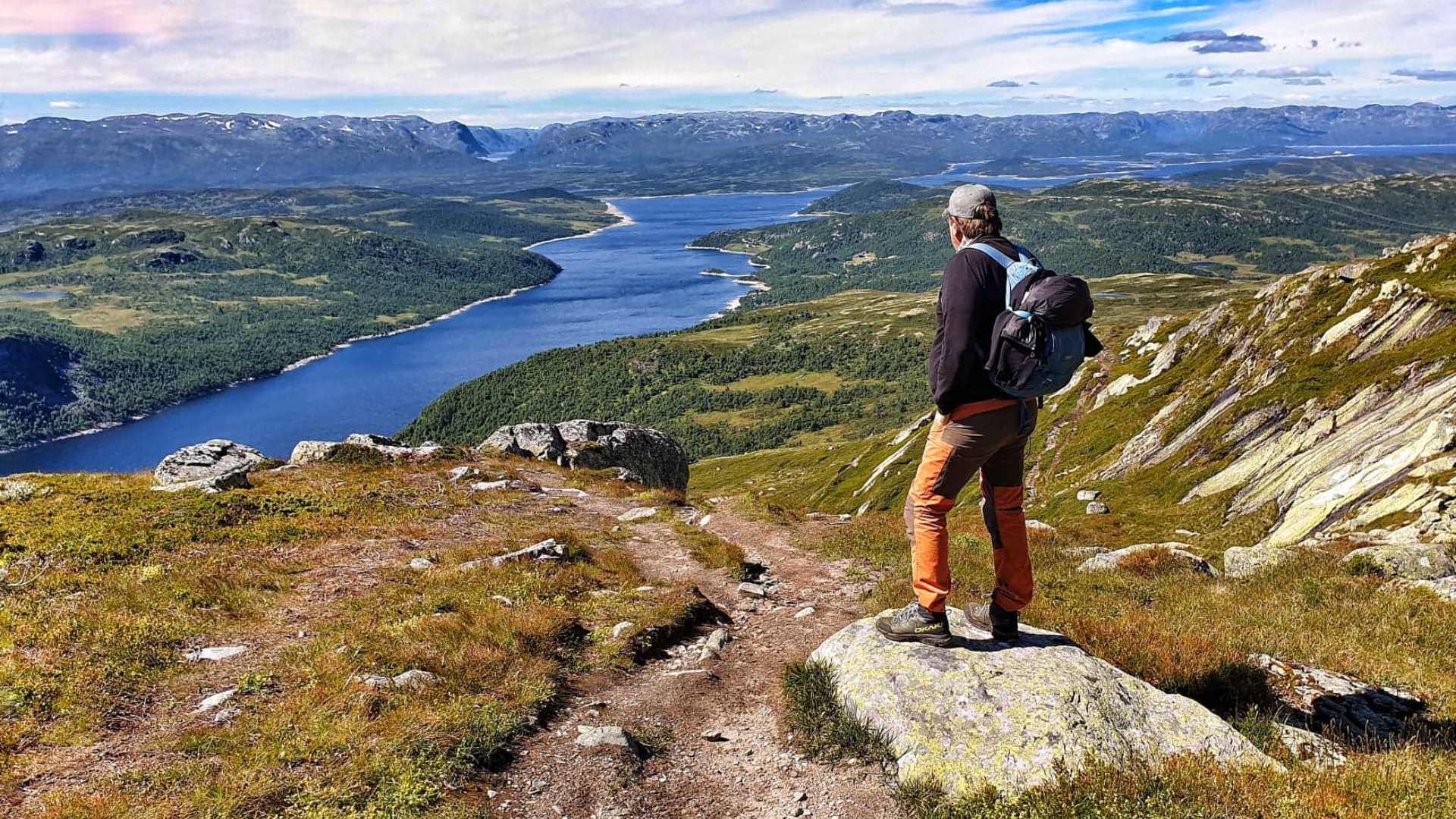 Vandrer ser utover fjord og fjell i Norge, med ryggsekk. Klar blå himmel og naturskjønn utsikt.