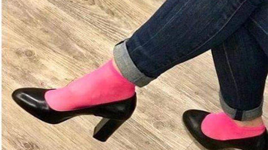 Person med rosa sokker i svarte høyhælte sko, iført oppbrettede jeans, på et tregulv.