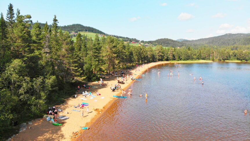 Strand ved innsjø omringet av skog og fjell, med mennesker som soler seg og bader i klart sommervær.