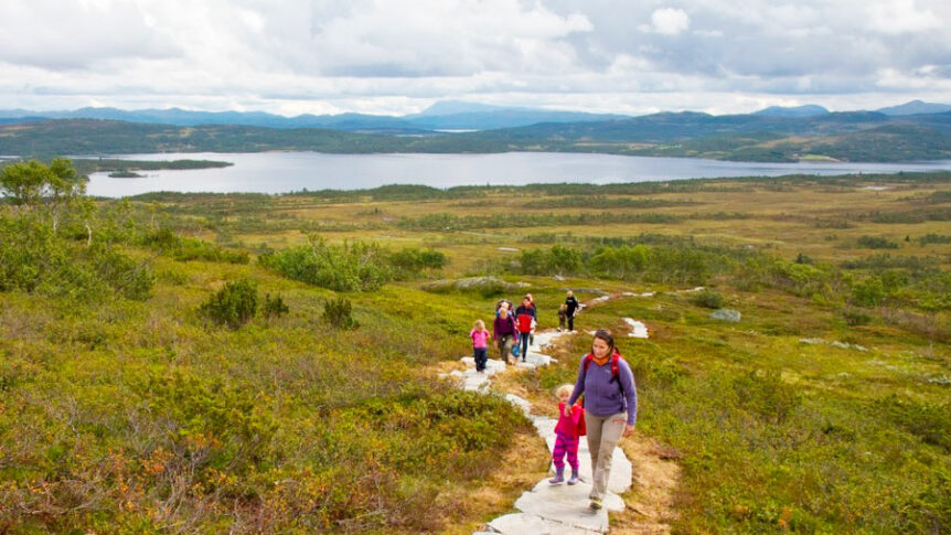 Familie på fottur i norsk fjellandskap med utsikt over innsjøen på en overskyet dag.