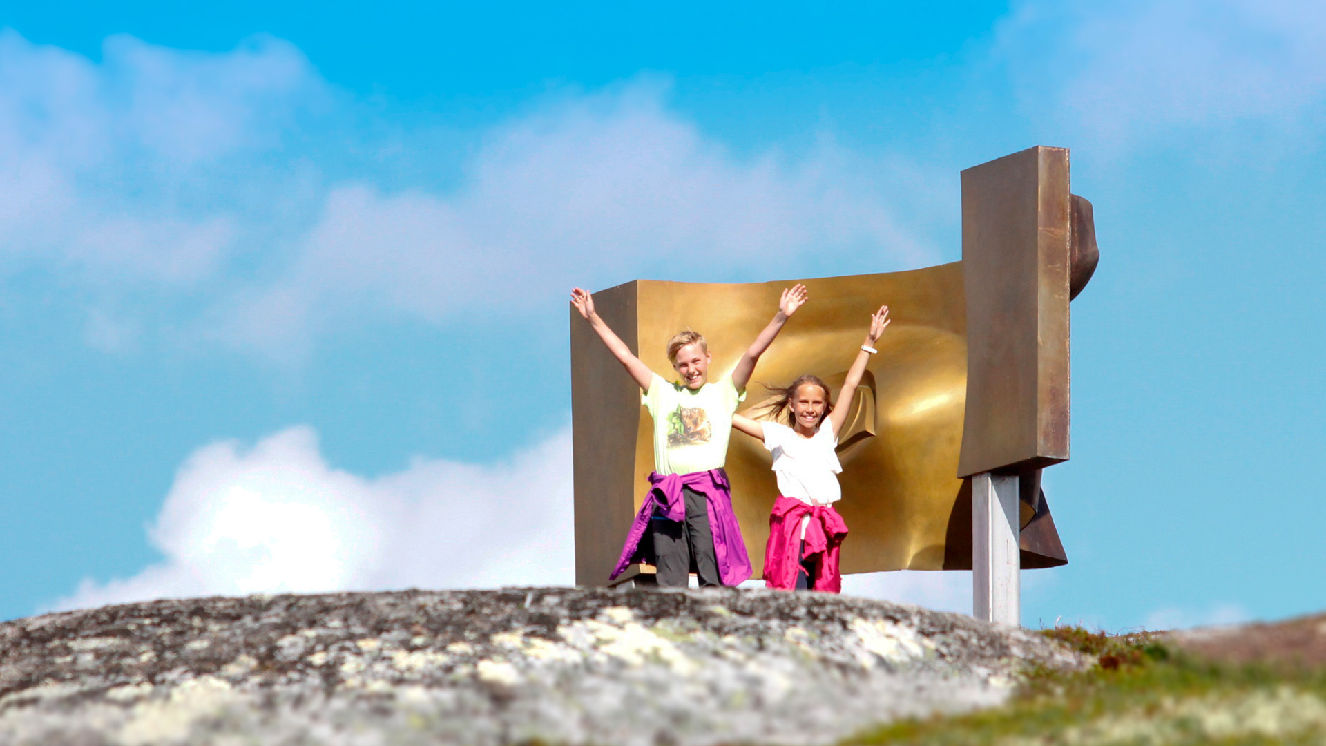 To barn på fjelltopp med skulptur i bakgrunnen, armene hevet under blå himmel.