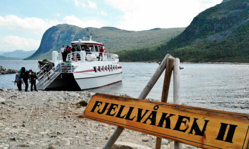 Fjellvåken II ferge ved innsjø omgitt av fjellandskap, passasjerer stiger av båten, solfylt dag.
