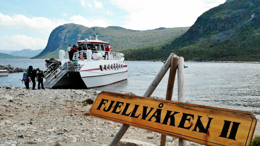 Fjellvåken II ferge ved innsjø omgitt av fjellandskap, passasjerer stiger av båten, solfylt dag.