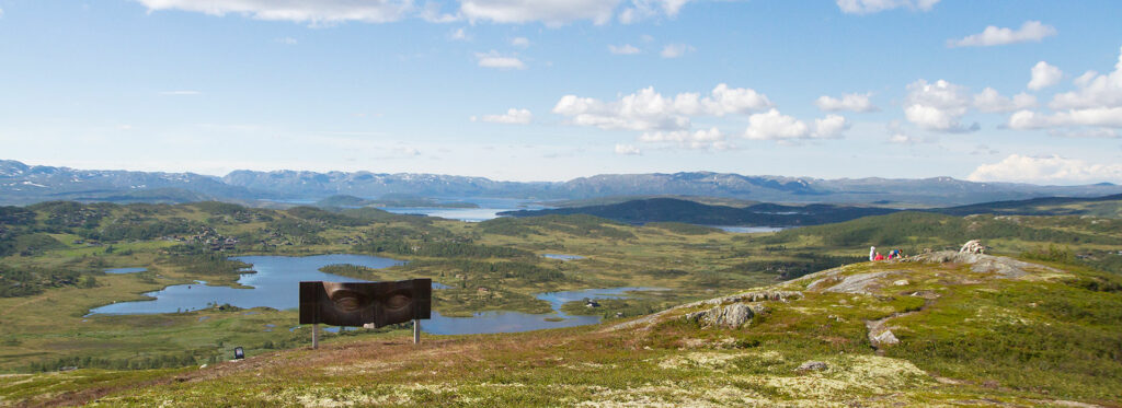 Utsikt over norsk fjellandskap med innsjøer, grønne åser og en kunstinstallasjon under blå himmel og skyer.