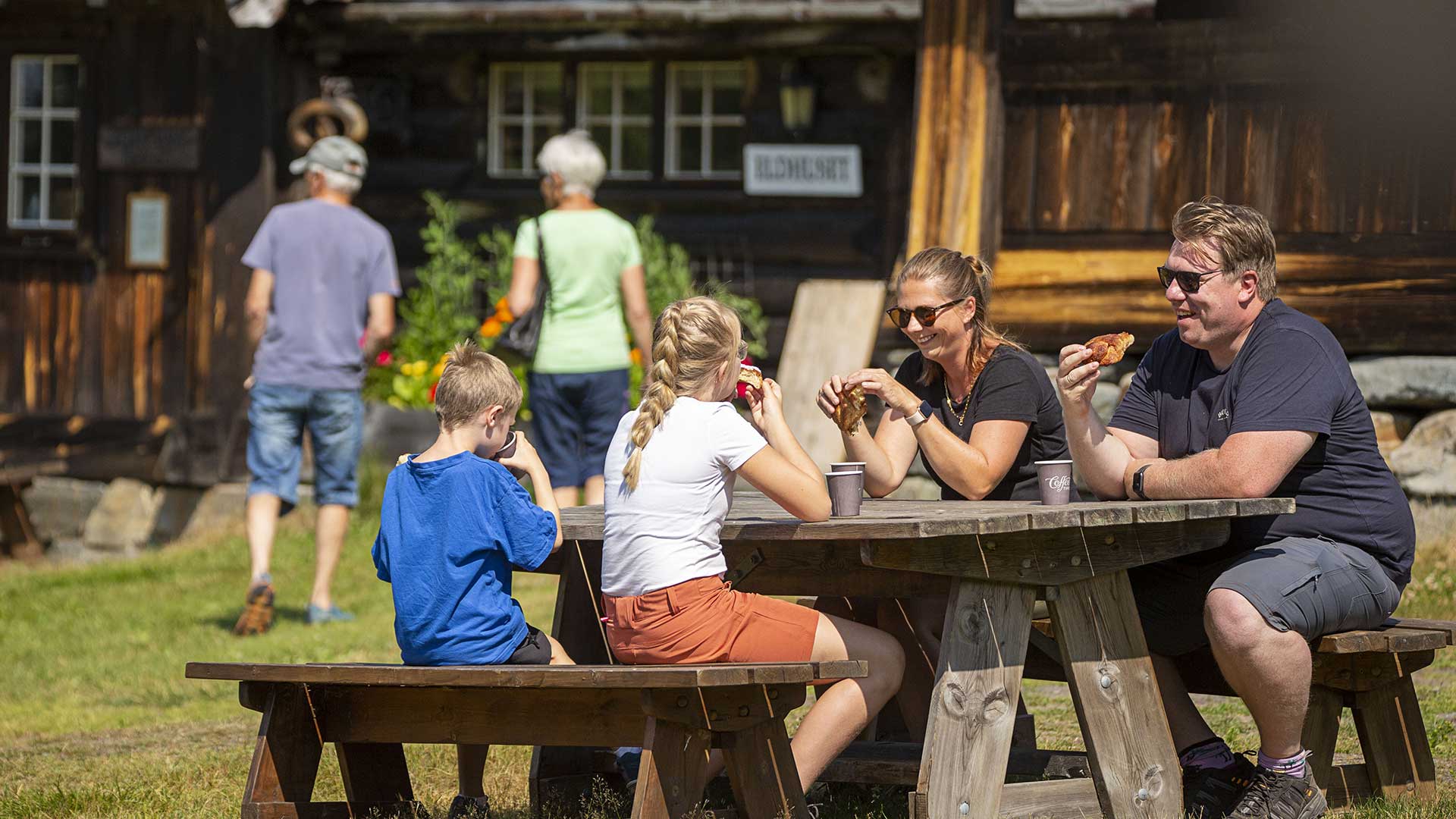 Familie nyter lunsj utendørs ved et bord foran en tradisjonell norsk hytte på en solrik dag.