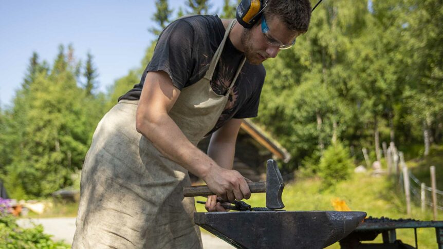 Smed arbeider utendørs med hammer og esse i en skogkledd setting.
