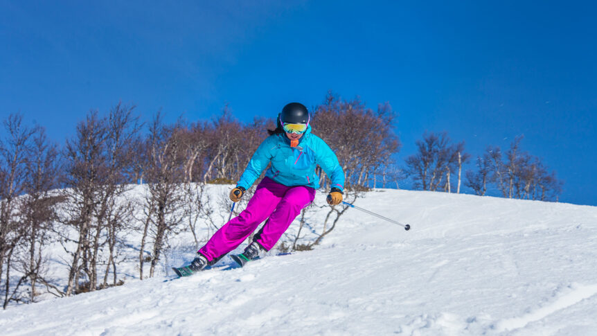 Person som står på ski ned snødekket bakke under blå himmel, iført turkis jakke og rosa bukser.
