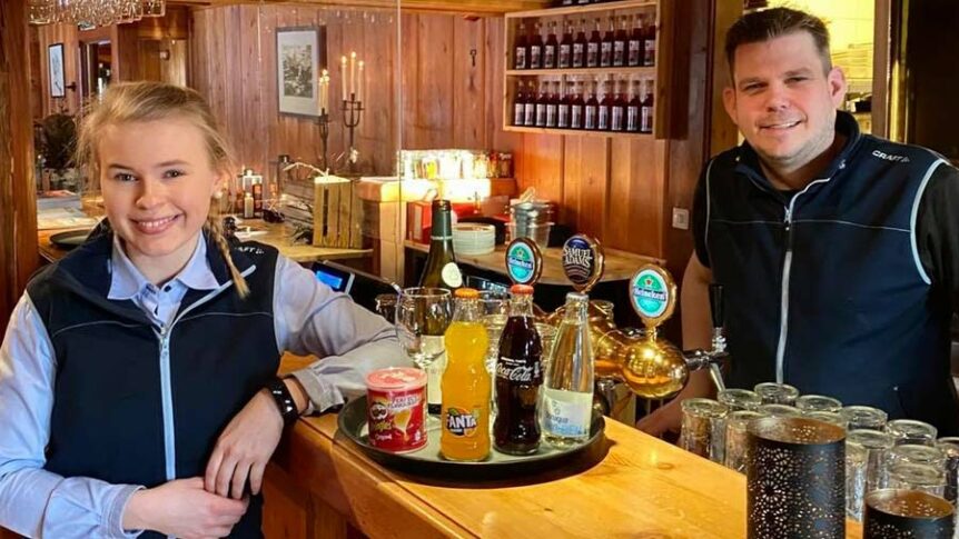 To bartendere i en koselig bar, omgitt av trepaneler og drikkevarer på disken.