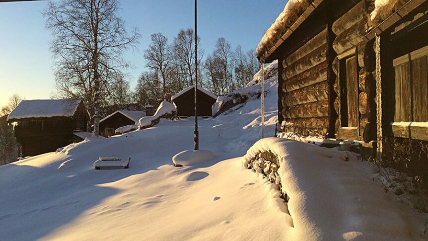 Snødekte trehytter i norsk landskap ved solnedgang, omringet av snødekte trær og klar himmel.