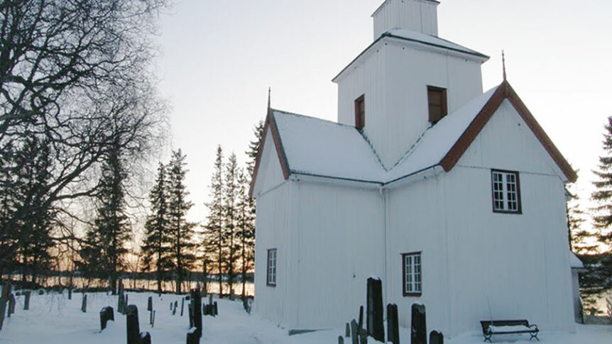 Hvit, snødekt trekirke med gravstøtter og skog i bakgrunnen. Vintersolnedgang skaper en vakker stemning.