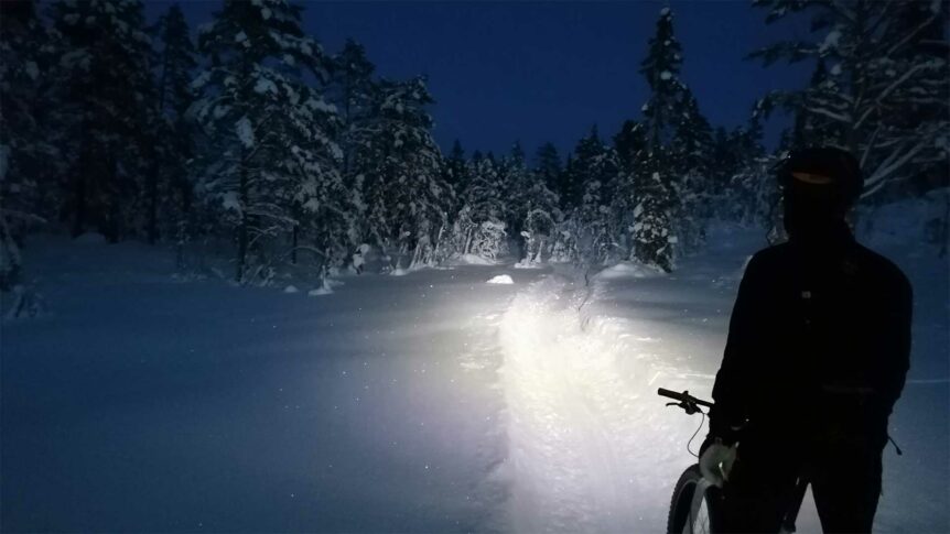 Sykkel på snødekt sti i skogen om natten, opplyst av frontlykt. Eventyrlig vinterlandskap.