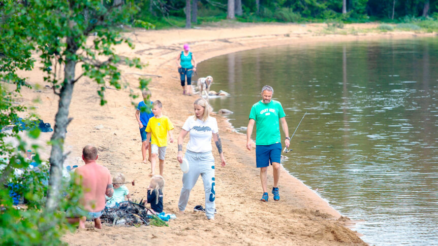 Familie som går tur på en strand ved en innsjø, med folk som fisker og leker, omgitt av natur.