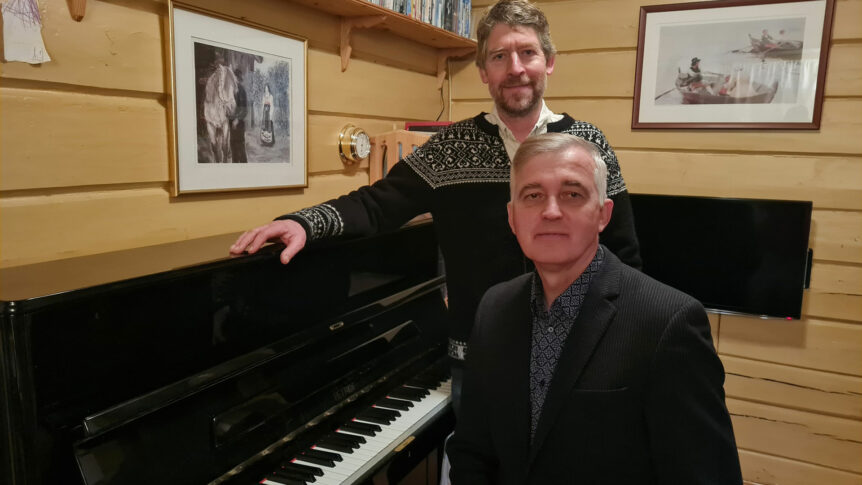 To menn ved et svart piano i en trehytte med bilder på veggen.