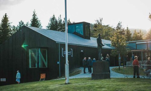 Besøkende ved et museum i Norge med skulptur og tradisjonell arkitektur i skogsområde.