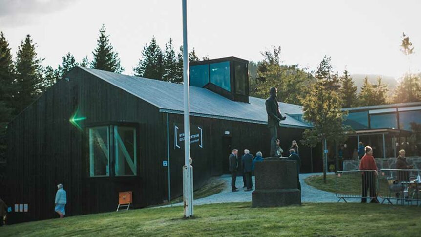 Besøkende ved et museum i Norge med skulptur og tradisjonell arkitektur i skogsområde.