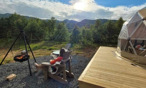 Person slapper av i stol ved bålplass, glamping-dom på treterrasse, omgitt av natur og fjellandskap under solfylt himmel.