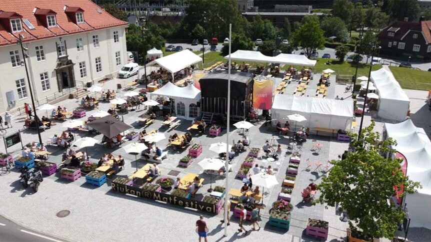 Luftfoto av Rjukan Matfestival med folk som nyter mat og drikke under parasoller på en solrik dag.