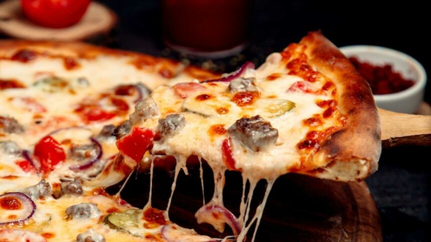 Nærbilde av en varm, gylden pizza med ost, tomat og løk, som blir løftet av en trespade.