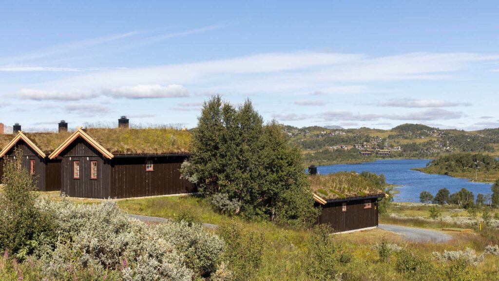 Tre hytter med torvtak ved et vakkert norsk landskap og blå innsjø under en klar himmel.