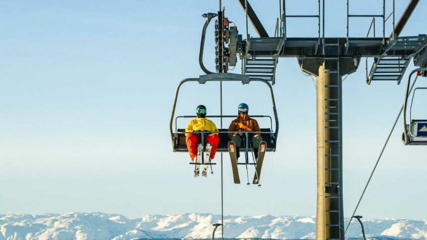 To personer på skitrekk med snødekte fjell i bakgrunnen. Klar blå himmel og vinterlandskap.