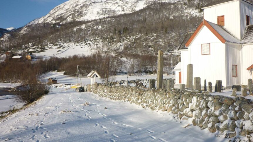 Vinterlandskap med snødekt fjell, kirkegård og hvitt hus i sollys i norsk dal.