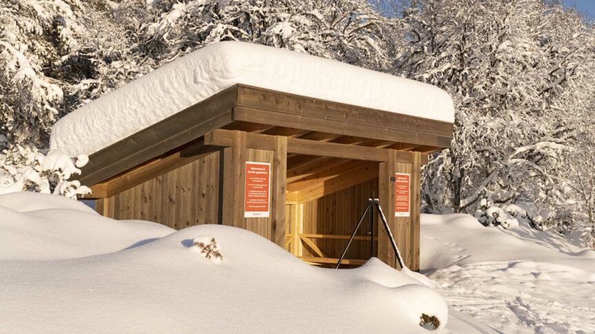 Snødekket gapahuk i skogen om vinteren, med trær og klart vær i bakgrunnen.