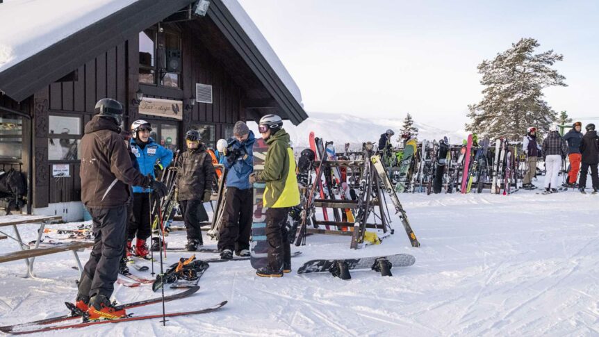 Skigjester samles utenfor fjellhytte med skiutleie og snowboardstativ. Vinterlandskap i fjellet.