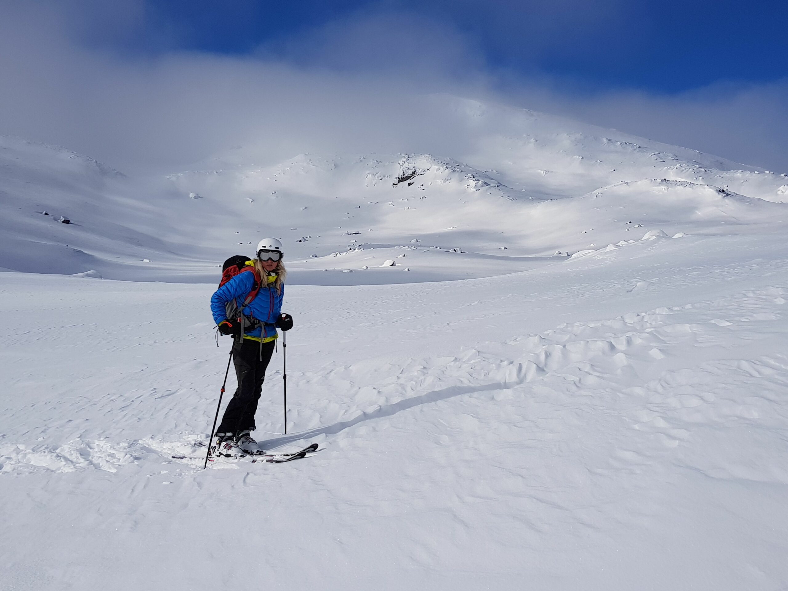En skiløper i blå jakke og hvit hjelm står på snødekte fjell med klar blå himmel i bakgrunnen.