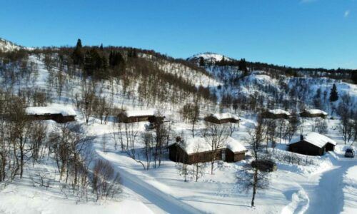 Snødekte norske hytter i fjellet under blå himmel, omgitt av vinterlandskap.