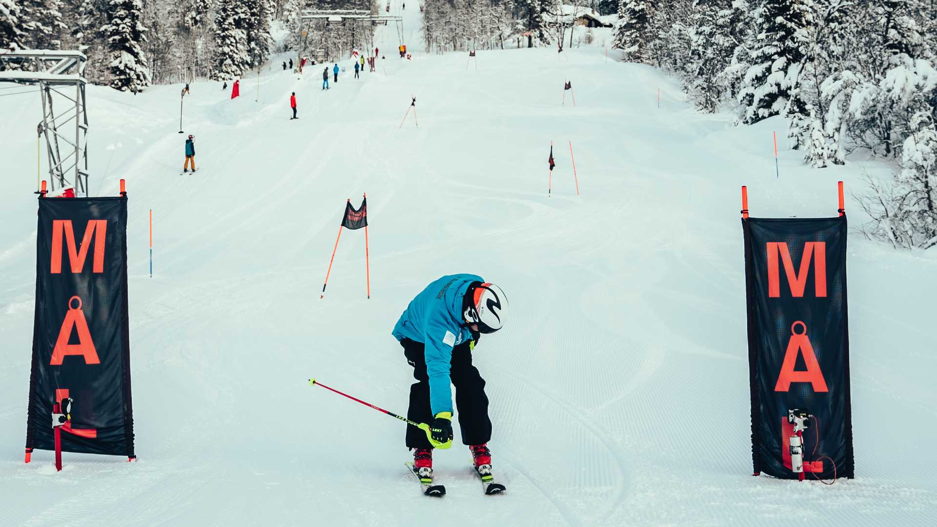Person iført skiutstyr ved målgang i skianlegg, omringet av snødekte trær og bakker.