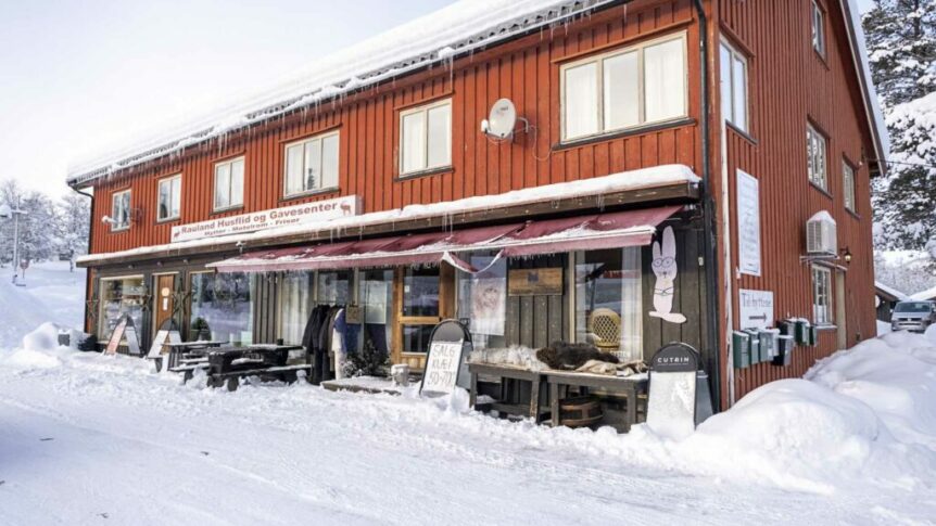 Rødt hus i vinterlandskap med skilt: Rauland Husflid og Gavesenter, omgitt av snø og butikkvarer utenfor.