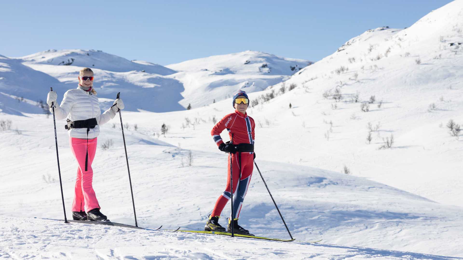 To personer på ski i solfylt vinterlandskap med snødekte fjell i bakgrunnen.
