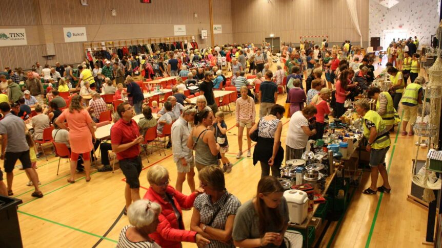Mange mennesker på en livlig loppemarked i en gymsal, med bord fulle av gjenstander og klær til salgs.