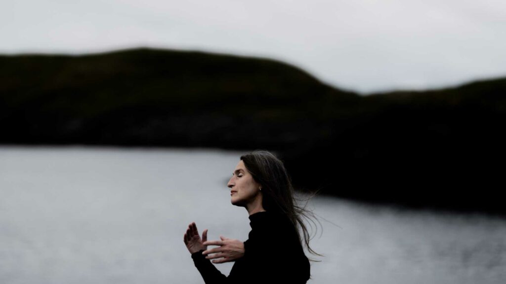 Kvinne ved sjøen i profil mot mørke fjell, i en meditativ positur med natur som bakgrunn.