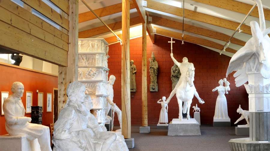 Utstilling av marmorstatuer i museum med naturlig lys fra takvinduer og røde murvegger i bakgrunnen.