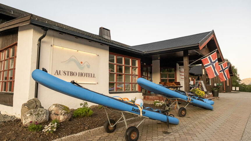 Utvendig fasade av Austbø Hotell med norske flagg og kajakker ved inngangen.