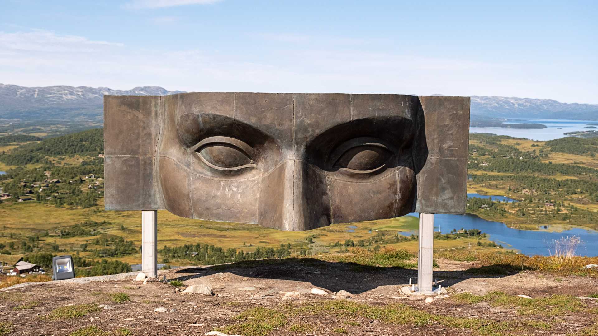 Skulptur på fjell med utsikt over dalen og fjorder i bakgrunnen, klar blå himmel, fokus på kunstverket.