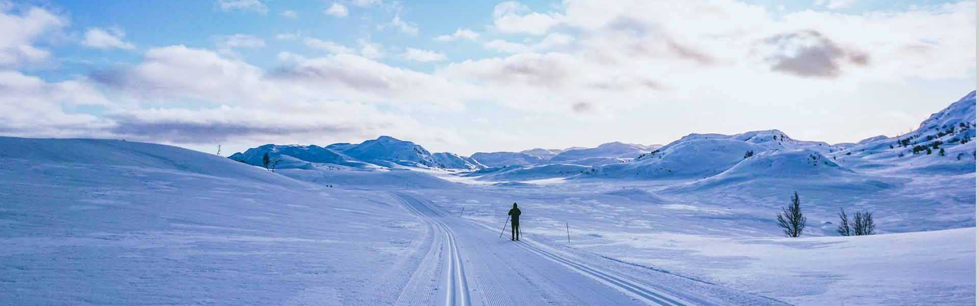 Person på langrenn i vinterlandskap med snødekte fjell og blå himmel.