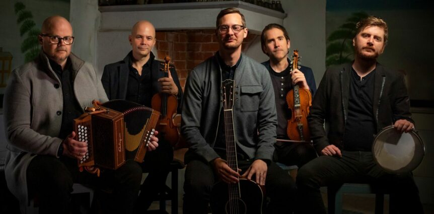 Fem musikere med ulike instrumenter, inkludert gitar og fele, sitter sammen i et rom.