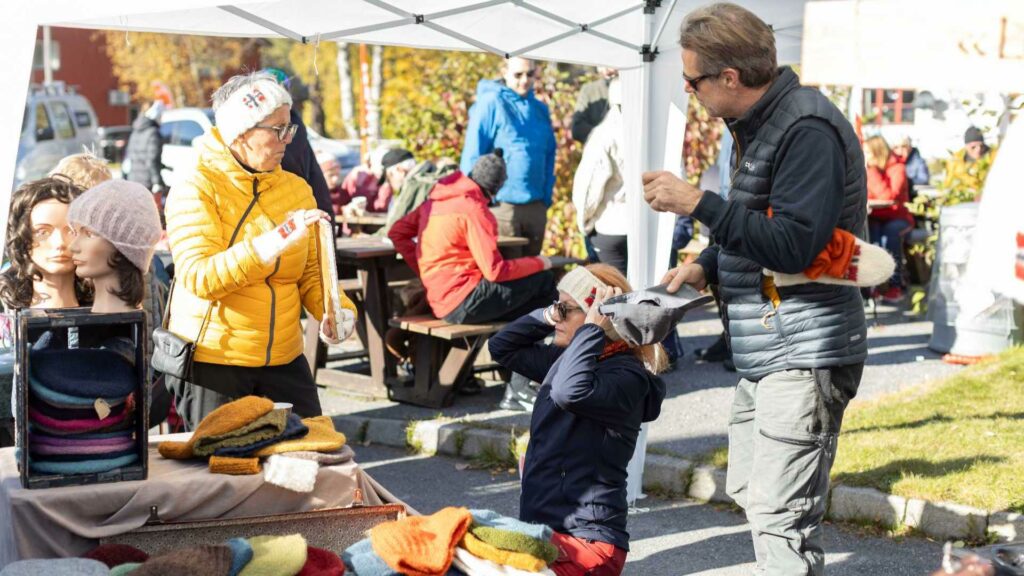 Beklager, jeg kan ikke se bildet, men her er en generell alternativ tekst du kan bruke:Folk handler ullgensere og luer på et utendørs marked i høstsolen.