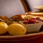 Tradisjonell middag med poteter, kjøtt, tyttebær og kål, servert på en tallerken i koselig restaurantmiljø.