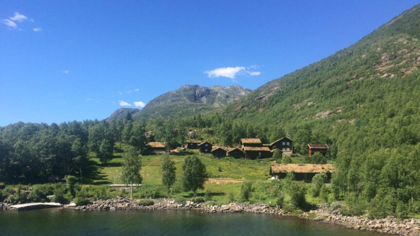 Norsk fjordlandskap med grønnkledde hytter, fjell og blå himmel i bakgrunnen.