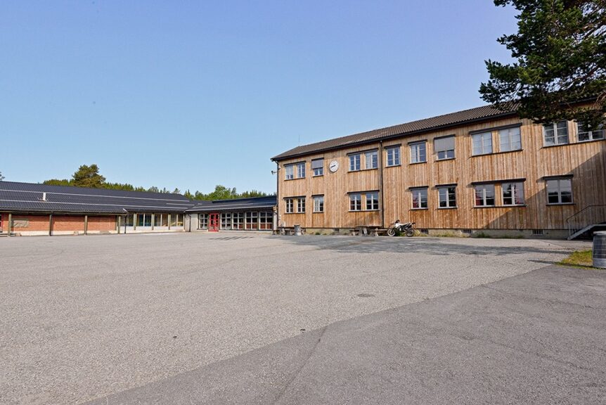 En tom skolegård med en stor trebygning under blå himmel, omgitt av trær.