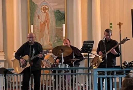 Band spiller musikk i kirke med gitar, trommer og bass foran religiøs kunst. Live opptreden i kirkerom.