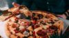 Person spiser en rykende varm pizza med ost, kjøtt og grønnsaker. Glass med drikke i bakgrunnen.