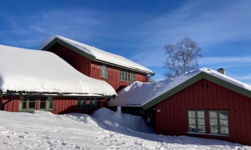 Rødt trehus med snødekt tak under blå himmel i norsk vinterlandskap.